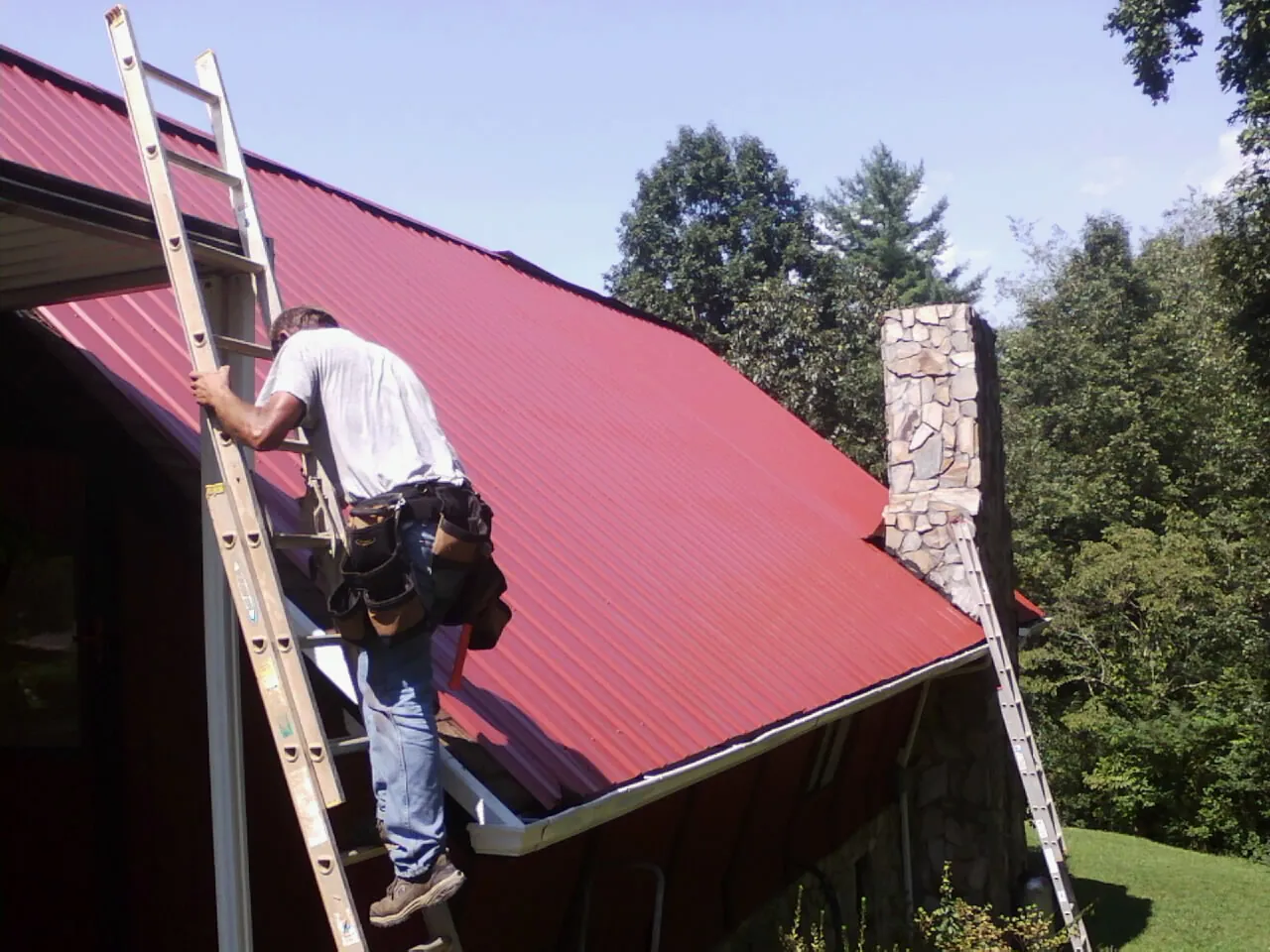 Roofer on a ladder beside a red metal roof and stone chimney — Soffit & Fascia Repair by Roofing Brothers in Maggie Valley
